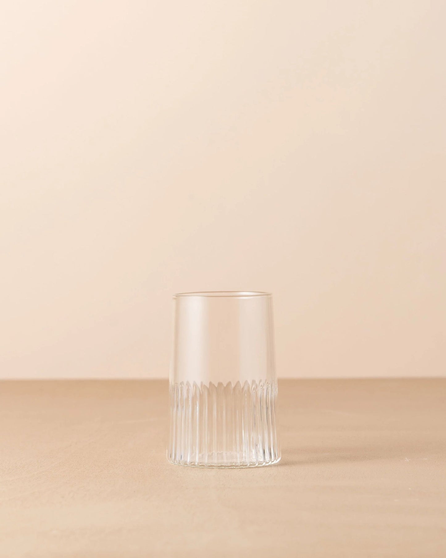 A KAIROS WATER GLASS by Saarde, featuring a clear and vertically ribbed design made of borosilicate glass, is displayed upside down on a smooth, neutral-toned surface against a light beige background. The simplicity and minimalistic style accentuate the texture and transparency of the glass.