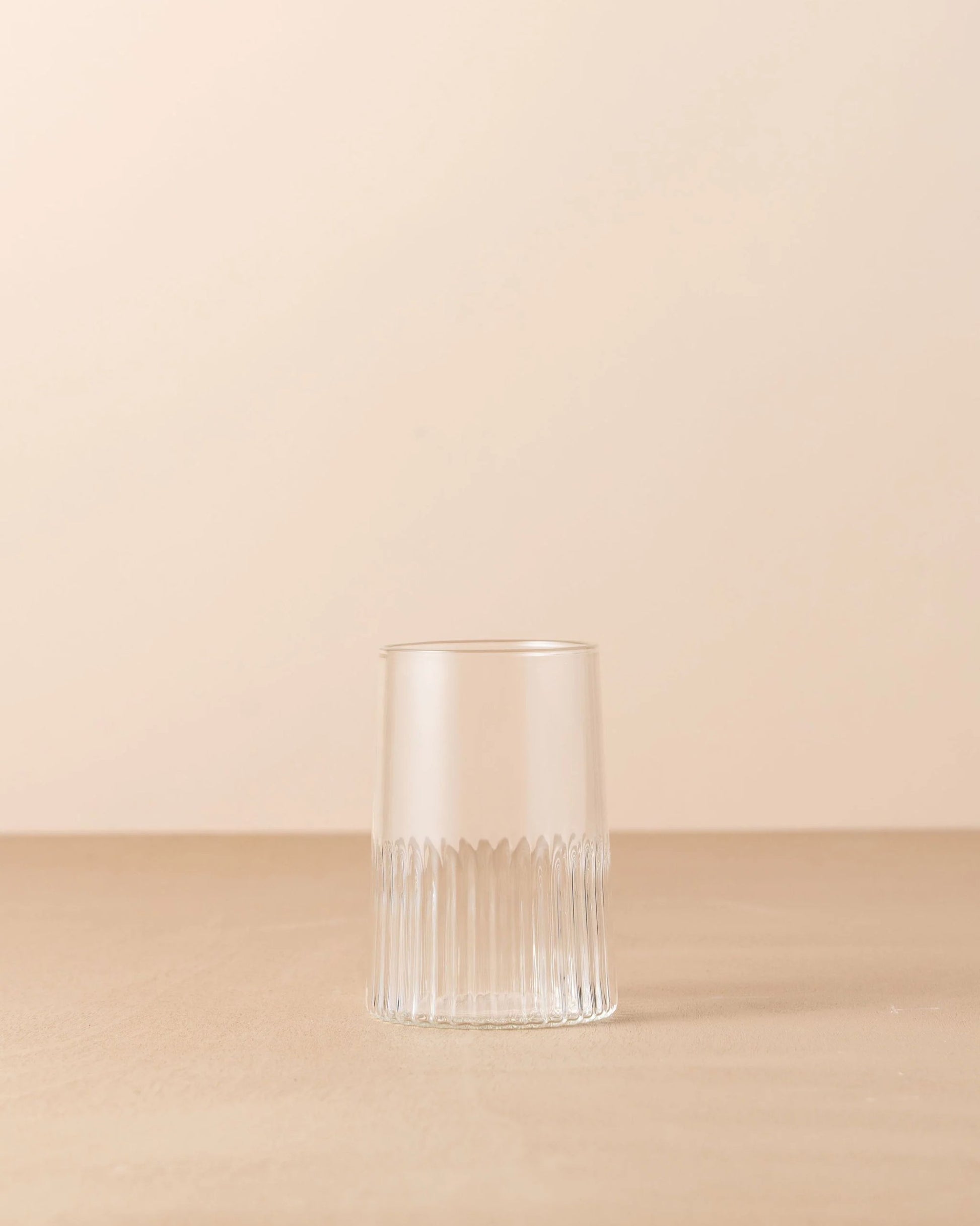 A KAIROS WATER GLASS by Saarde, featuring a clear and vertically ribbed design made of borosilicate glass, is displayed upside down on a smooth, neutral-toned surface against a light beige background. The simplicity and minimalistic style accentuate the texture and transparency of the glass.