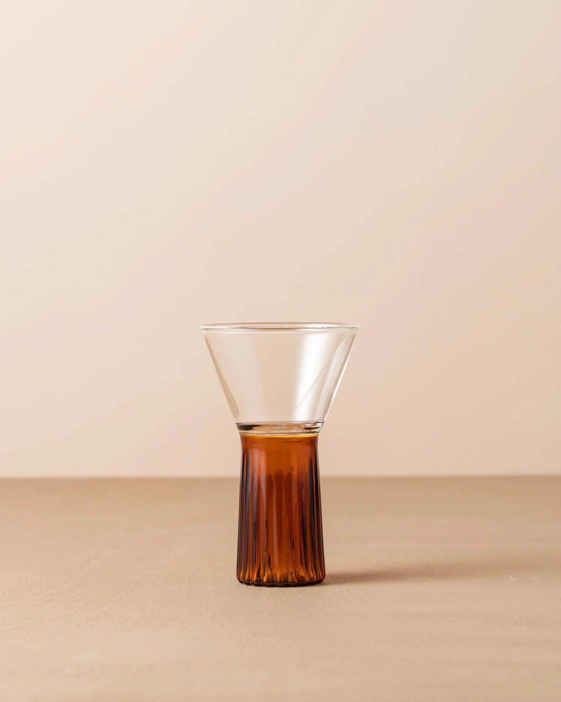 The KAIROS WINE GLASS by Saarde, featuring a clear glass design with an amber base, rests on a beige surface against a light tan background. Evoking the charm of vintage Murano glassware, this elegant piece has a ribbed texture in its lower section and transitions to a smooth conical shape at the top.