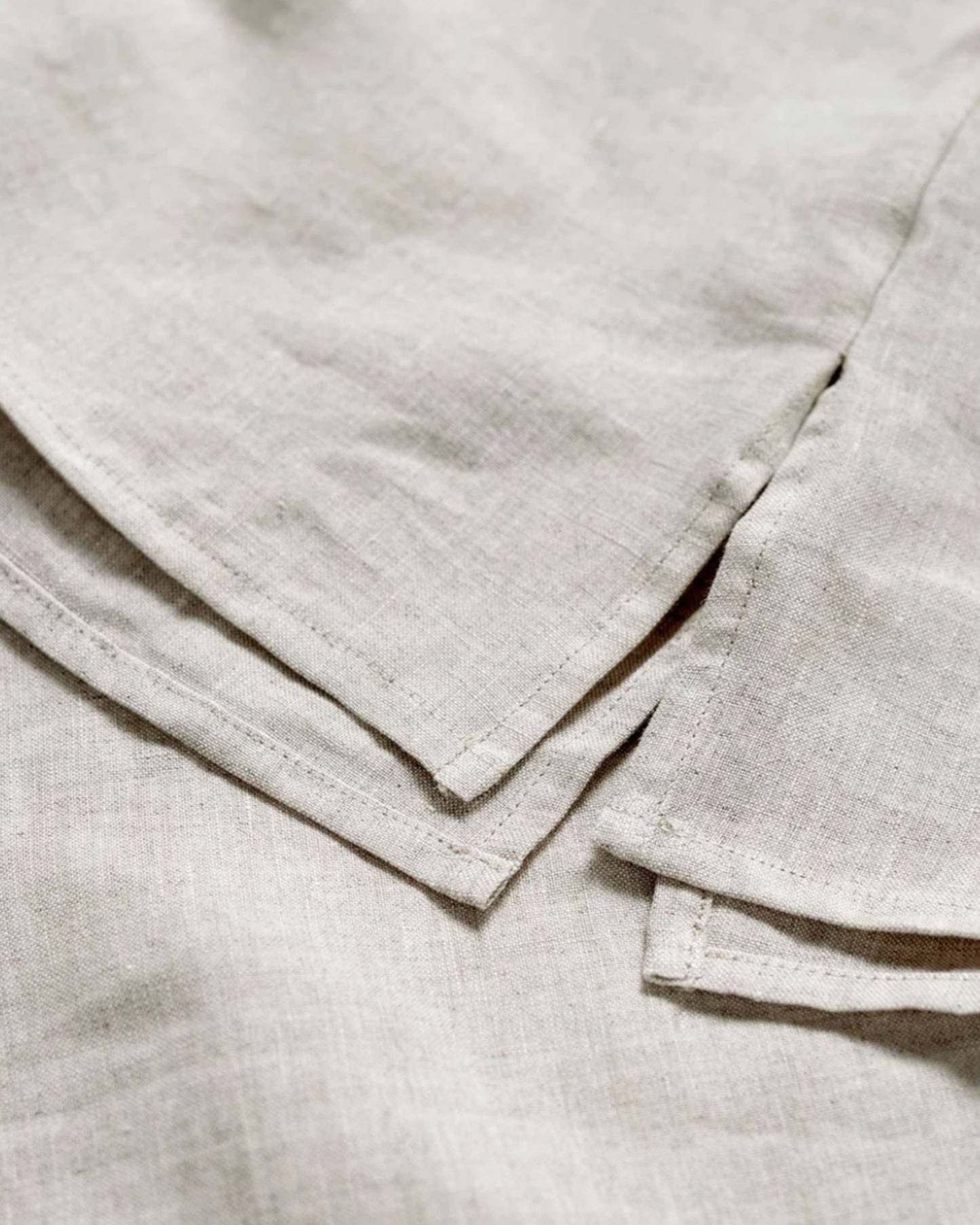 A close-up of the LE LIN VALANCE by The Workroom showcases a light beige fabric with visible texture and overlapping edges. Reminiscent of French flax linen, the material appears soft and woven, with neatly folded corners. This image captures the simplicity and detail of this earthy-toned textile.