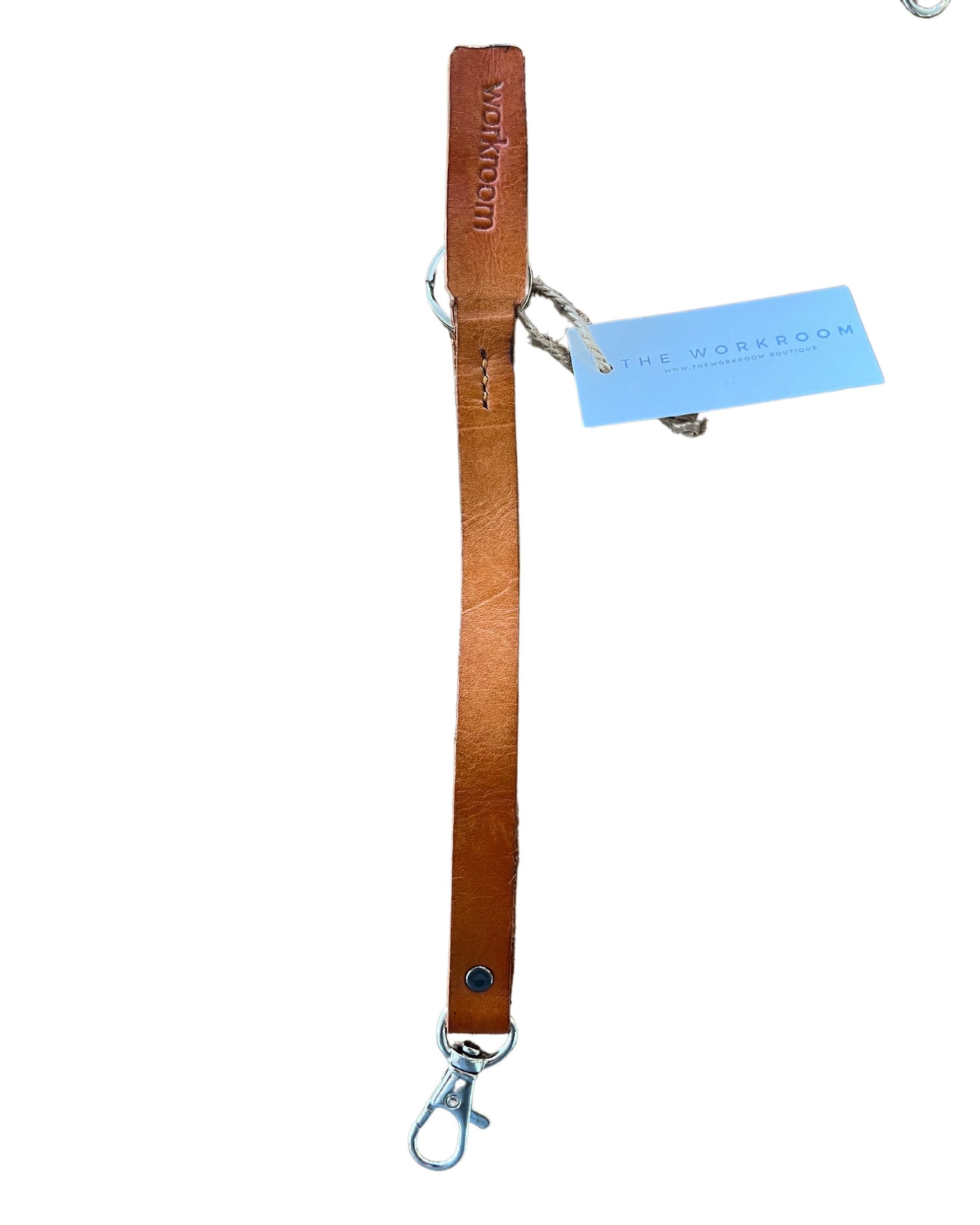 The LEATHER KEY CHAIN from The Workroom boasts a tan full-grain leather wrist strap, complete with a metal clasp and branded tag for an enduring charm. The strap is embossed with "Workroom" near the top. A light blue card inscribed with "The Workroom" is attached using twine, combining style and practicality in one elegant piece.