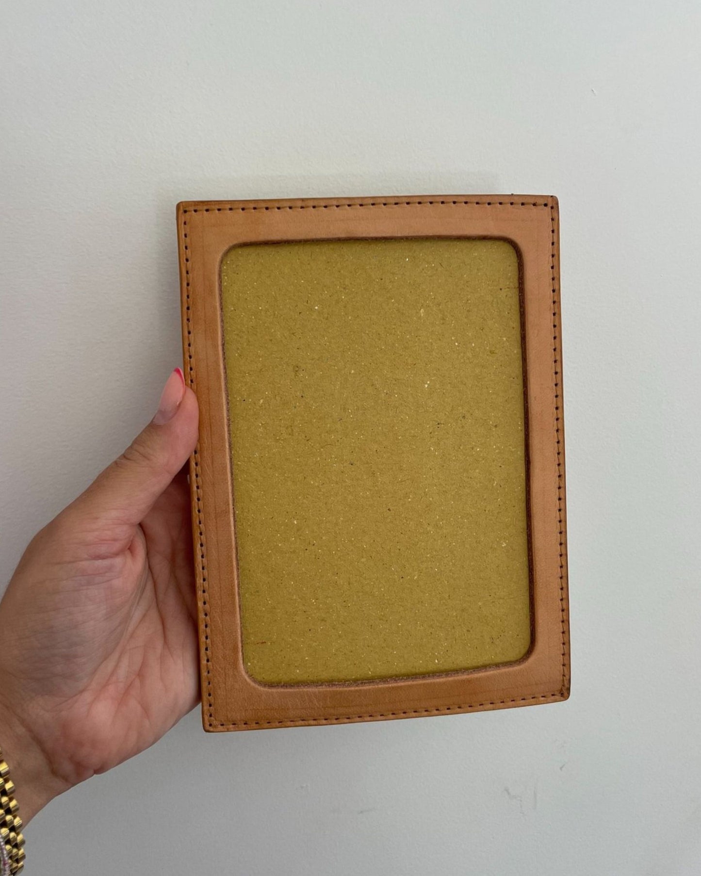 A hand is holding The Workroom's LEATHER PHOTO FRAME, featuring a minimalist design in tan leather, against a plain white background.