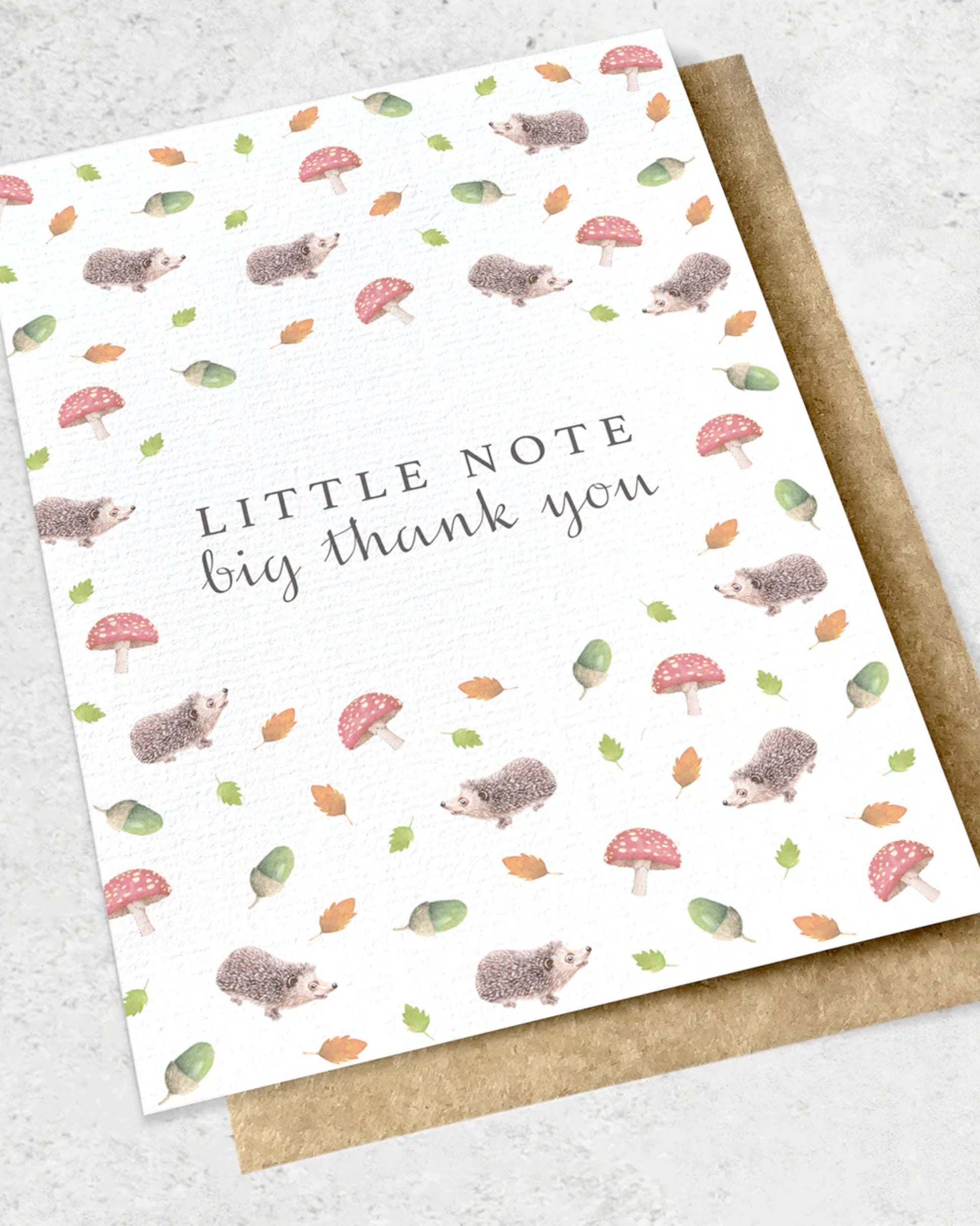 Displayed on a gray surface is the Ink Bomb "LITTLE NOTE BIG THANK YOU - GIFT CARD," featuring hedgehogs, mushrooms, acorns, and leaves in soft colors. A kraft envelope peeks out from beneath.