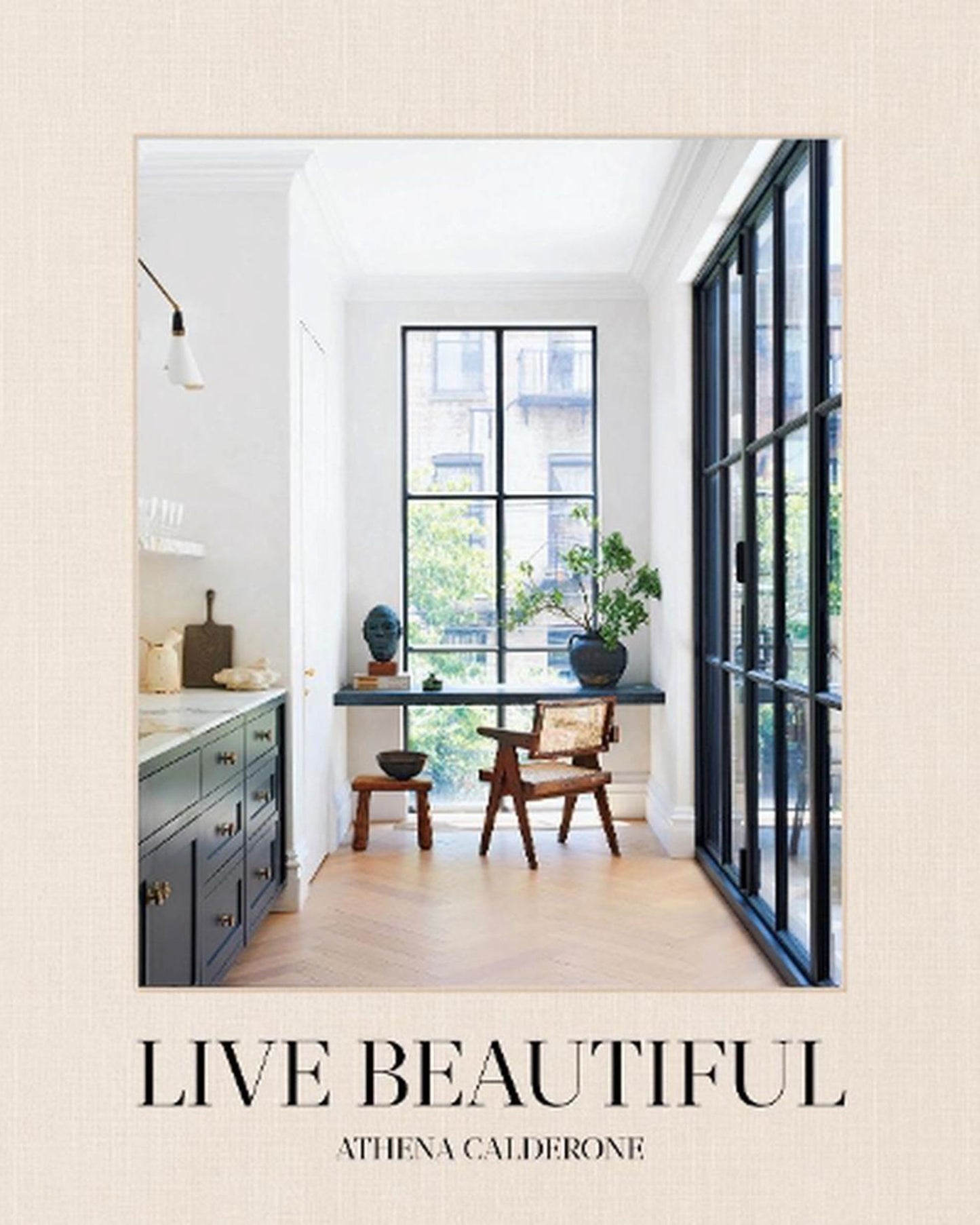 The cover of "Live Beautiful - Athena Calderone" by Nation Wide Books showcases a bright and elegant interior featuring large windows, a small table with two chairs, and plants. This design book includes stunning photography that captures a modern, minimalist style perfect for interior decorators.