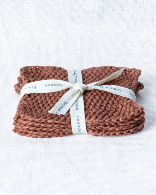 A brown, knitted washcloth crafted from organic cotton is neatly folded and tied with an off-white ribbon embossed with the brand name Bianca Lorenne. This sustainable choice, ideal as a reusable makeup remover called Lavette Makeup Remover, rests on a textured white surface.