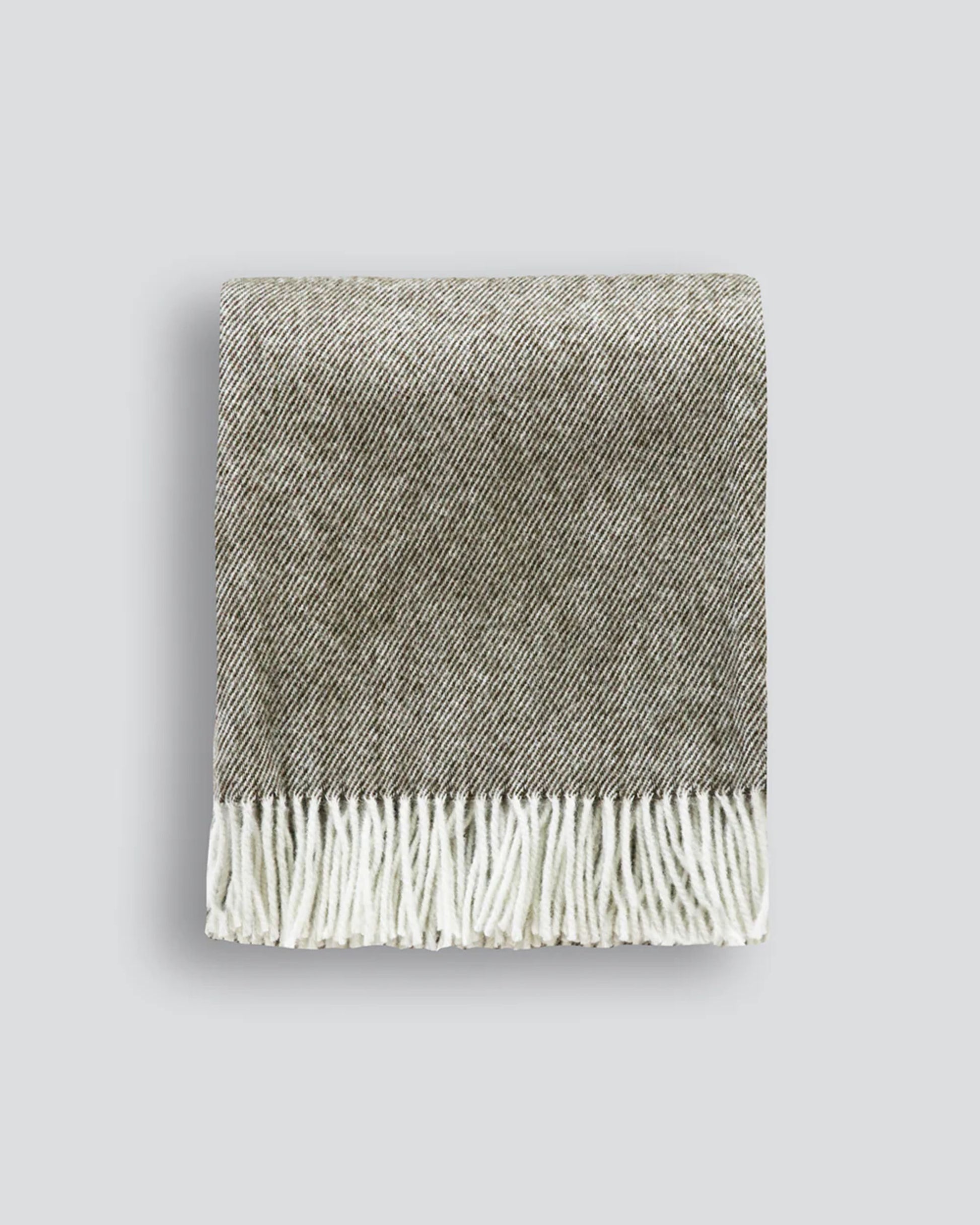 The MARTINBOROUGH THROW from Baya, a luxurious folded gray wool blanket adorned with white fringes, rests on a light gray background. Locally crafted in Hawke’s Bay, it showcases a subtle herringbone pattern for an elegant touch.