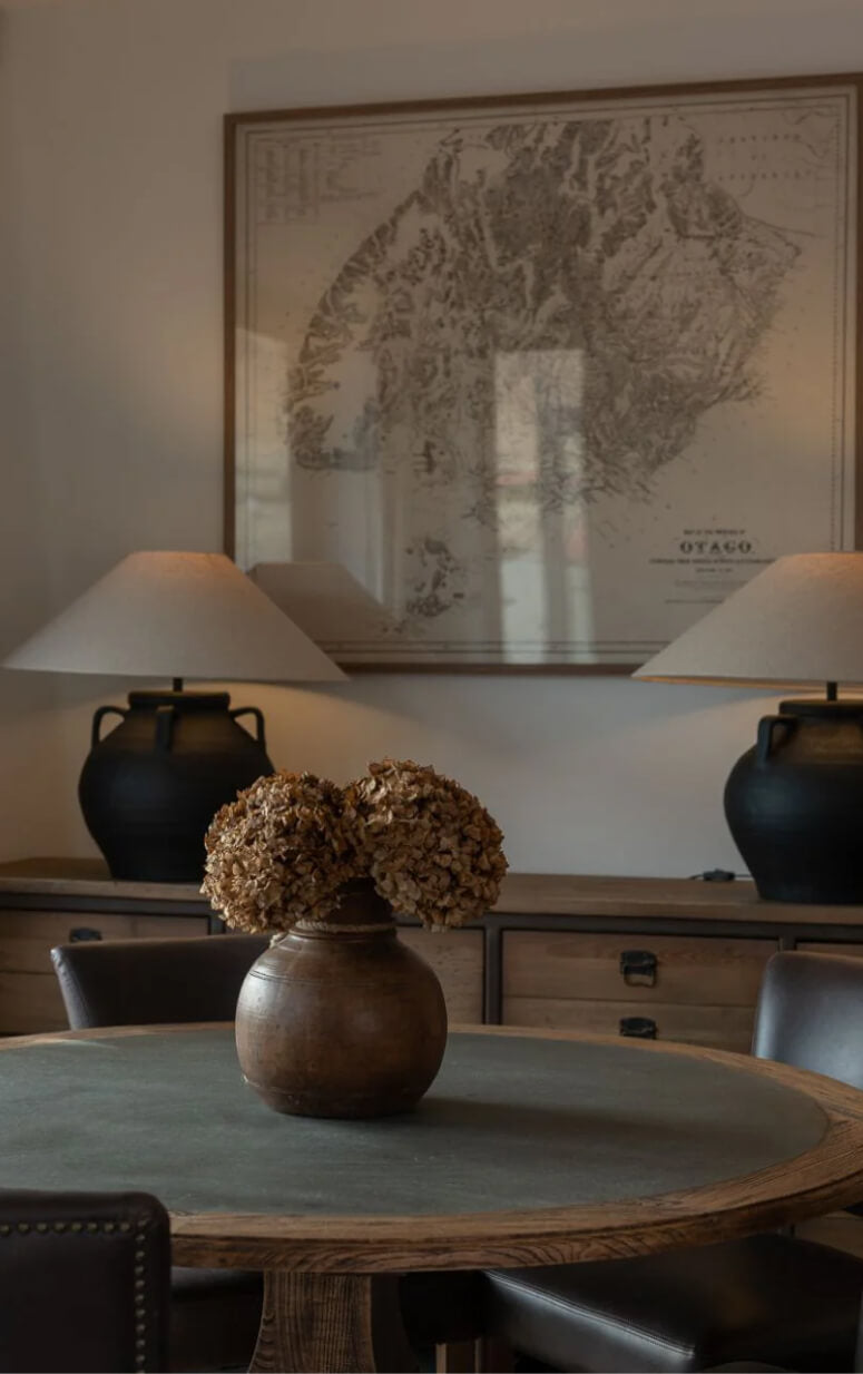 A cozy room with a round table featuring a vase of dried flowers. Two black lamps with beige shades sit on a wooden console against the wall. A large framed antique map hangs behind, adding a vintage touch to the decor.