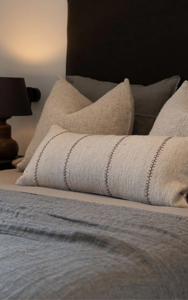 A cozy bedroom setup with a neatly made bed featuring gray and beige pillows. A beige lumbar pillow with a stitched pattern is at the front. Soft lighting is provided by a black lamp on a wooden bedside table.