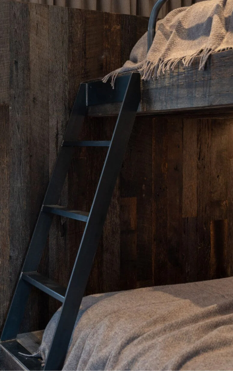 A cozy wooden bunk bed with a dark, rustic finish. The bottom bunk is covered with a gray blanket, while a ladder leads up to the top bunk, also draped with a gray blanket. Warm lighting enhances the inviting atmosphere.