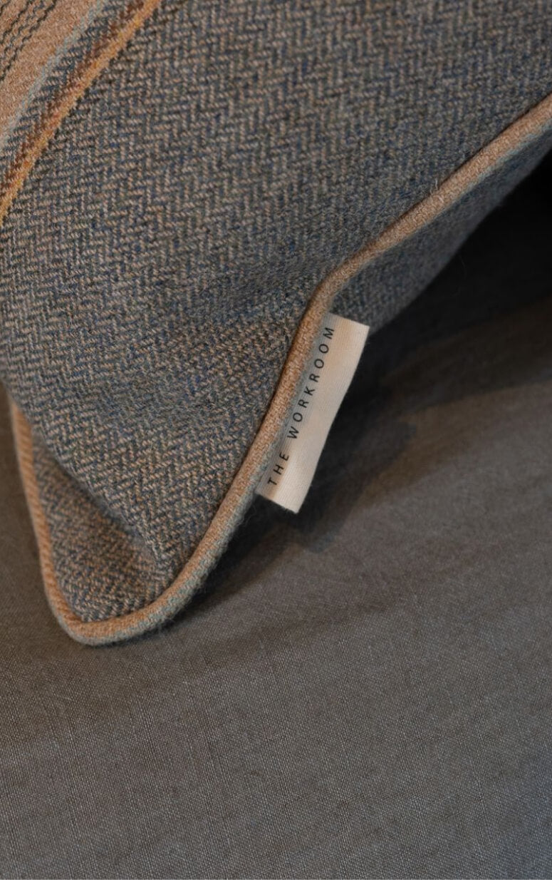 A close-up of a textured gray pillow with brown trim and stripe detail. A small tag reading THE WORKROOM is visible on the side. The pillow rests on a complementary gray fabric surface.