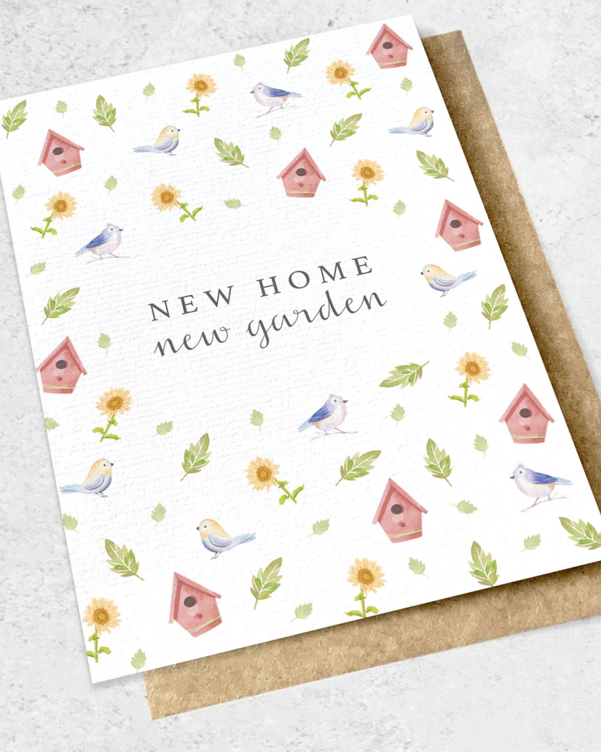 Introducing the Ink Bomb "NEW HOME NEW GARDEN" Greeting Card: a premium textured card adorned with birds, birdhouses, sunflowers, and leaves. The phrase "New Home New Garden" elegantly decorates the center. It comes with a kraft envelope and is set against a light background, perfect for impressing any recipient.