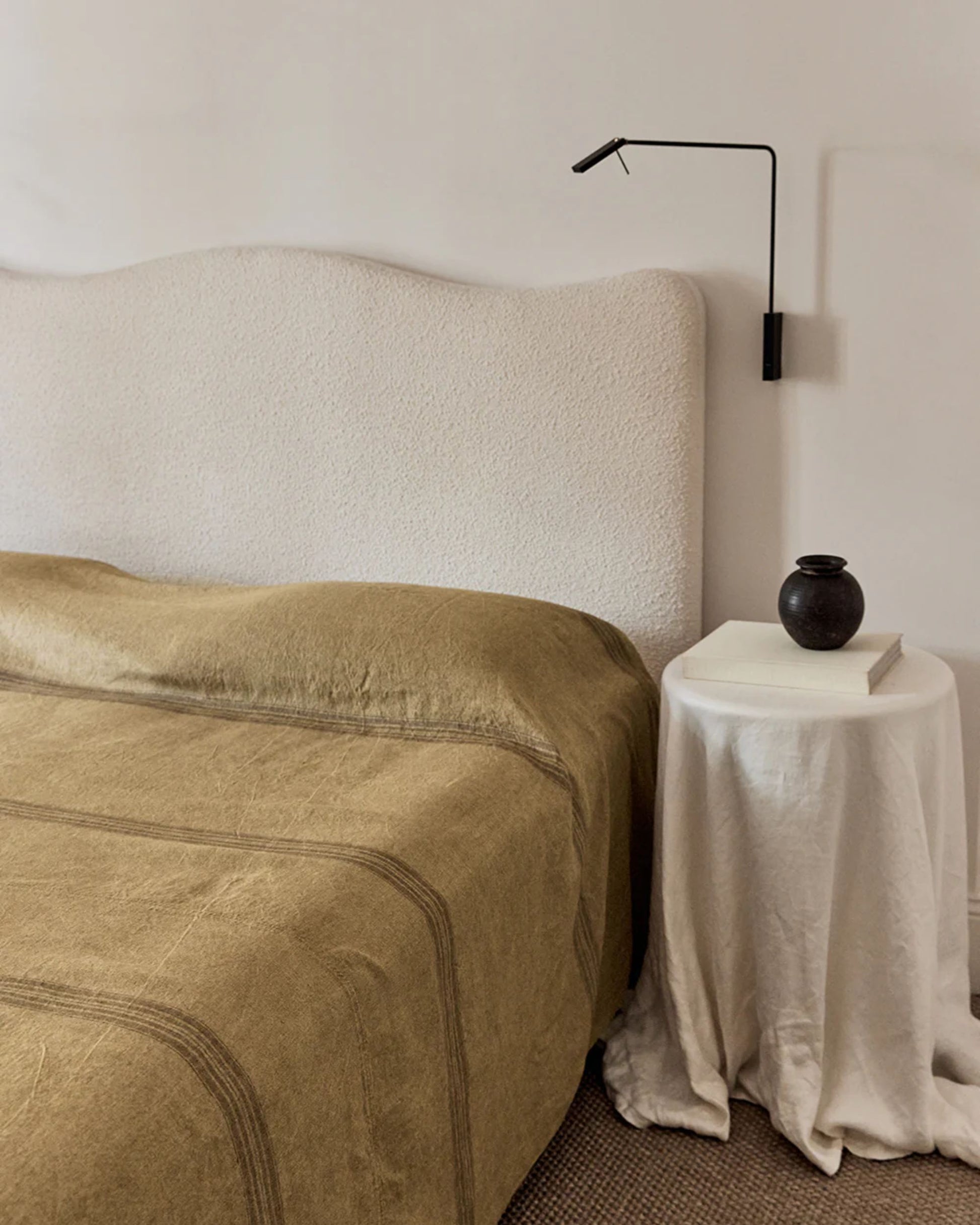 In a minimalist bedroom, an In the Sac NOMAD BED COVERLET in textured beige and 400gsm linen takes center stage, accompanied by a cream cushioned headboard. A small side table with an elegant drape displays a black vase and book, softly lit by a sleek black wall lamp above.