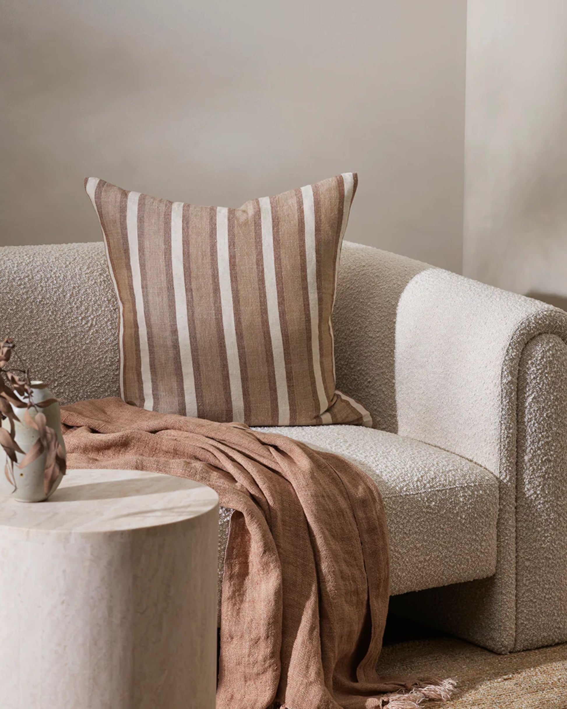 A cozy living room scene with a textured beige sofa, featuring a handwoven Baya Brighton cushion. A soft brown throw blanket made of natural fibers is draped over the arm. In the foreground, a round coffee table with a decorative vase completes the arrangement.