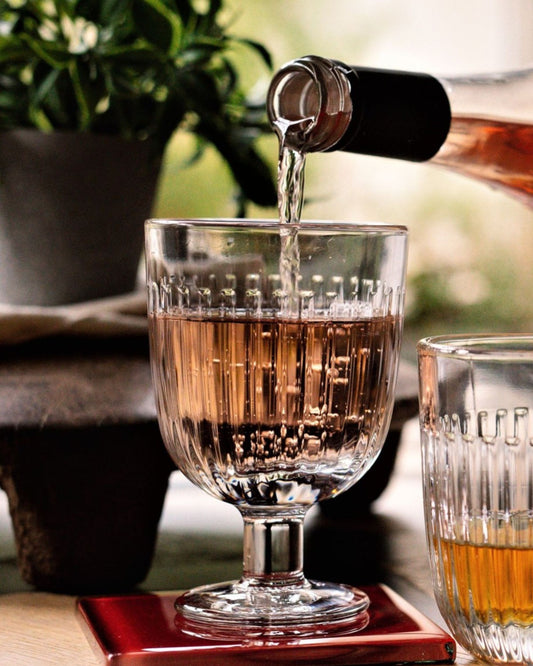 A close-up of rosé wine being poured into an OUESSANT WINE GLASS by La Rochere, known for its ornate design and dishwasher-safe convenience, on a table. Nearby is another glass containing a different drink. The background displays a blurred green plant.