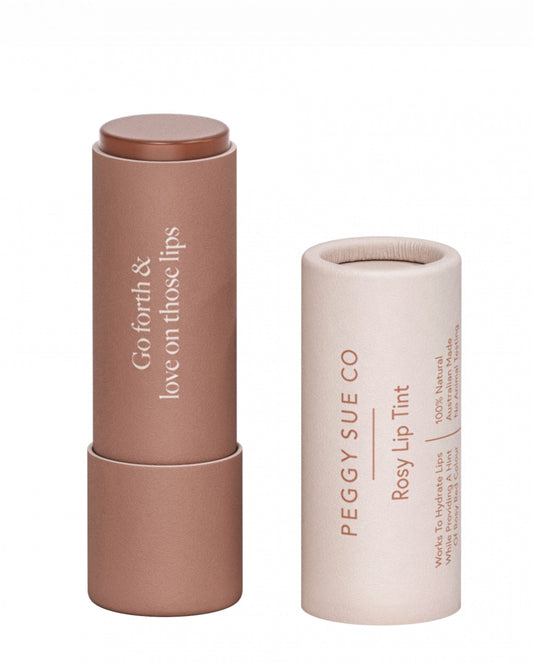 A rosy lip tint called PEGGY SUE - ROSEY LIP TINT by Peggy Sue is displayed in a pinkish-brown, cylindrical tube alongside its matching cap. The tube is infused with natural ingredients and features the phrase "Go forth & love on those lips," while the cap is labeled with "Peggy Sue Co Rosy Lip Tint," making it ideal for maintaining a healthy hydration barrier.