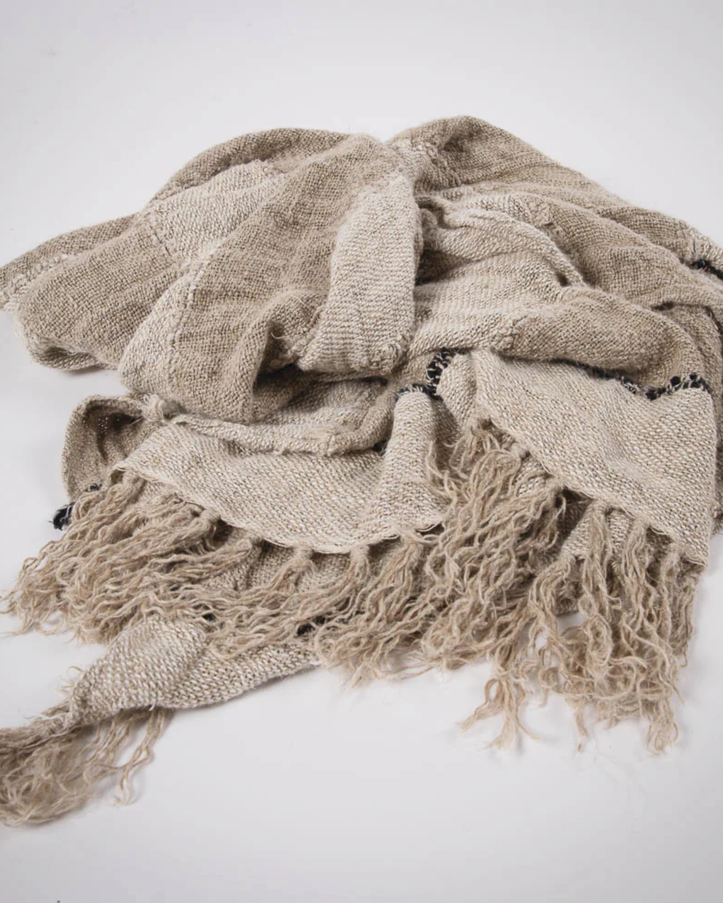 A crumpled RETREAT THROW in natural, ivory, and black hues from Eadie lies on a white surface, highlighting its cotton/linen blend. The fabric looks textured and soft, with a natural earthy tone that captures the rustic charm of the throw.