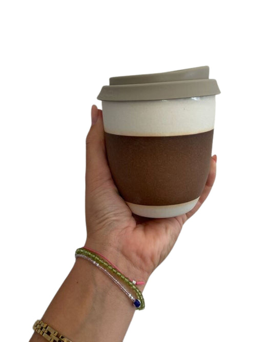A hand adorned with a colorful bracelet holds the WORKROOM REUSABLE CUP by Workroom, featuring a beige lid and brown band, expertly crafted from Australian stoneware clay in a Noosa studio, against a plain white background.