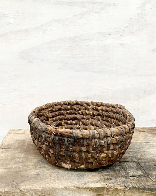 The FRENCH ANTIQUE HAND-COILED RYE BREAD BASKET by All Things French rests on a wooden surface against a simple, light backdrop. Its textured weaving and varied shades of brown impart a rustic, artisanal charm.