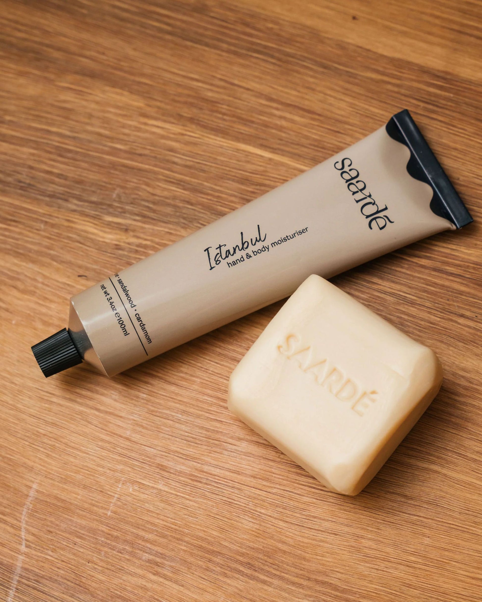 A beige tube of Saarde HAND AND BODY MOISTURISER, infused with sandalwood, is placed on a wooden surface alongside a beige soap bar featuring the engraved Saarde logo.