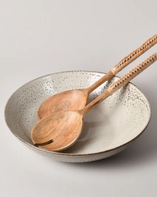 A pair of "SALAD SERVER W/RATTAN HANDLE" by New Direction rests on a speckled ceramic bowl, which exudes a natural, earthy appearance against a plain background.