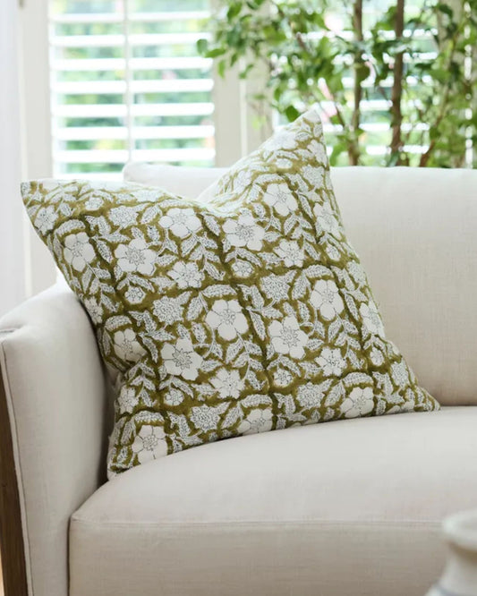 A cream-colored sofa is adorned with the SALTA FLORAL CHARTREUSE CUSHION COVER and placed near a bright window with white blinds. The soft linen enhances the room's decor, while a lush green plant adds an elegant touch in the background.