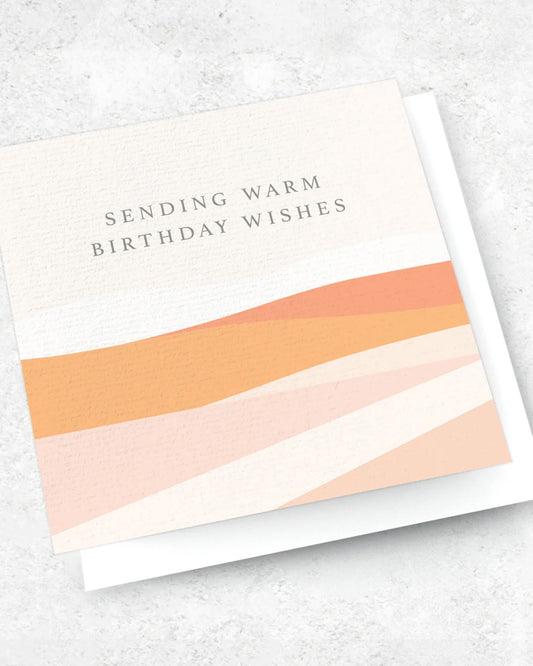 Introducing the SENDING WARM BIRTHDAY WISHES - GREETING CARD by Ink Bomb, a premium greeting card featuring the elegantly scripted message "Sending Warm Birthday Wishes" on the front. The design beautifully displays abstract wavy patterns in soft shades of orange, peach, and cream. It comes complete with an envelope and is presented resting on a light gray surface.