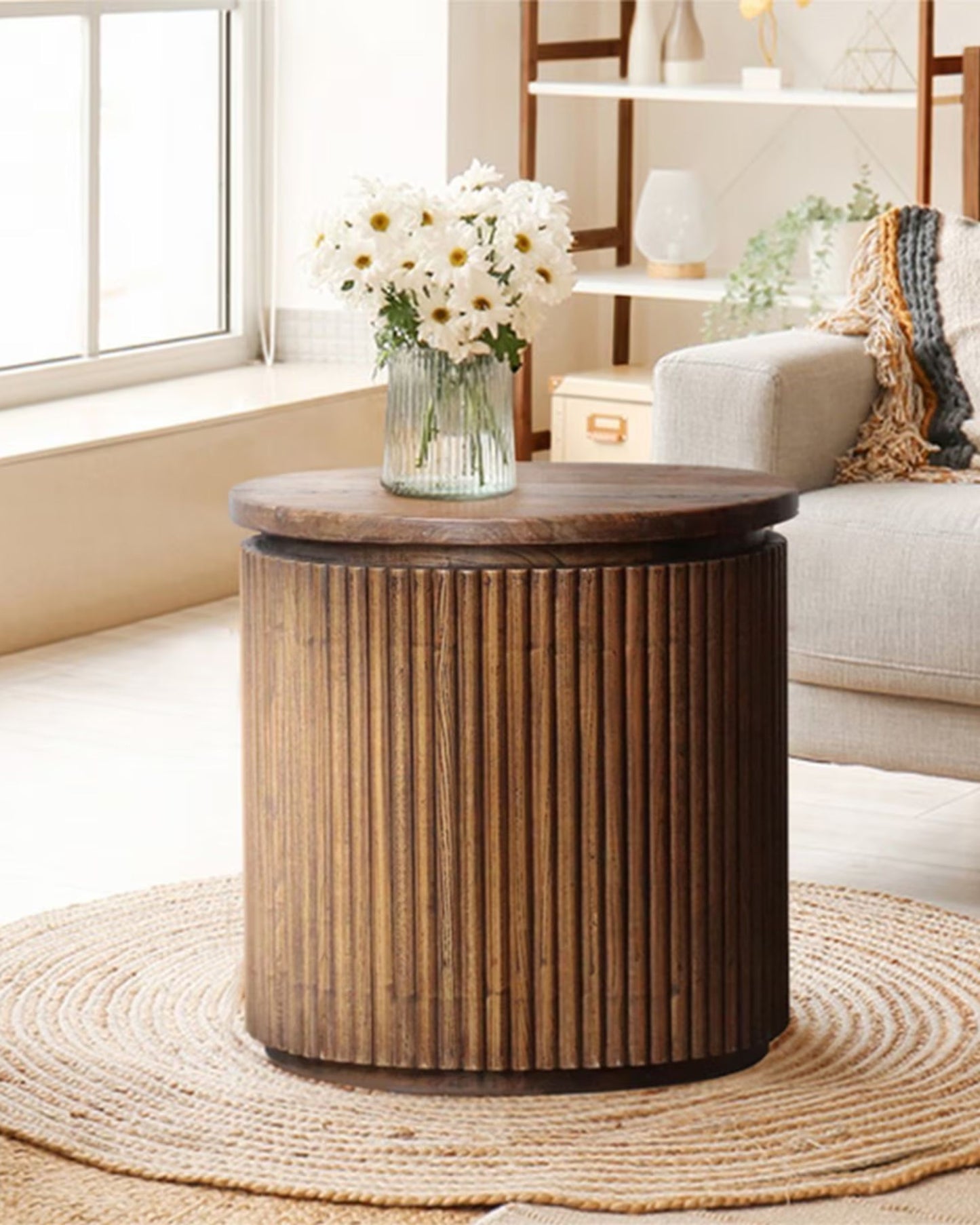 A SIDE ROUND TABLE - OLD ELM by Rembrandt with vertical slats is set on a textured rug, exemplifying contemporary design. A glass vase with white flowers rests on its surface. Close by, there's a gray sofa and a shelf adorned with decor items. Sunlight flows through a large window, offering a cozy atmosphere.