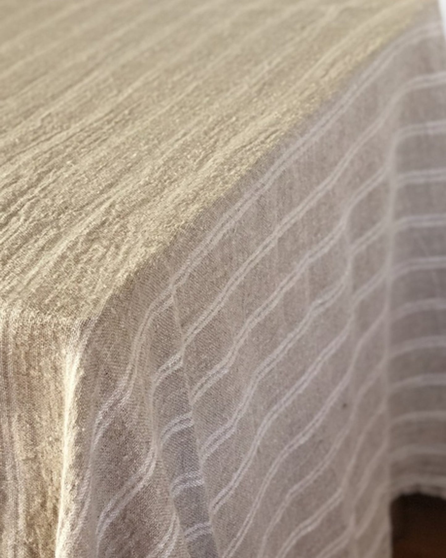 A close-up of a white, handloomed tablecloth with a striped pattern draped over a table evokes an elegant look. This textured, woven piece adds charm to any home decor, much like the Rustic Linen Bed Throw/Table Cloth, known for its sophisticated design.