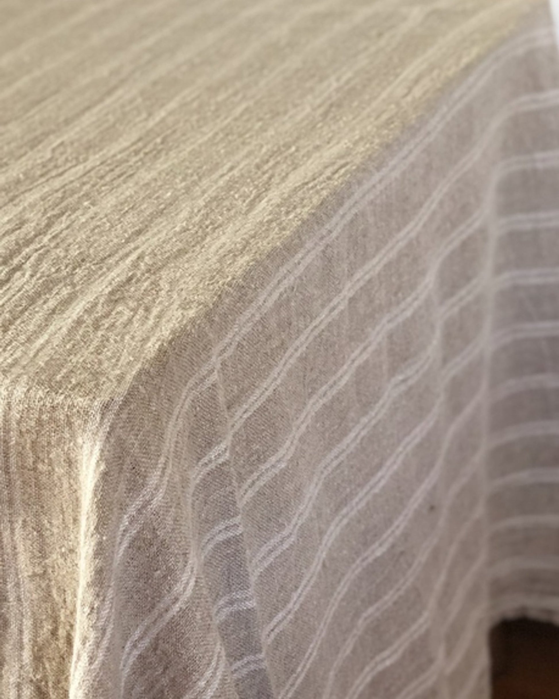 A close-up of a white, handloomed tablecloth with a striped pattern draped over a table evokes an elegant look. This textured, woven piece adds charm to any home decor, much like the Rustic Linen Bed Throw/Table Cloth, known for its sophisticated design.