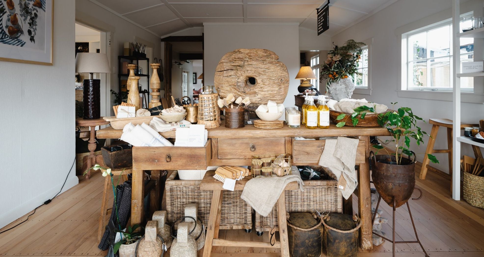 A cozy, rustic interior showcasing a wooden table with various home decor items, such as baskets, candles, and bottles. The space is bright, with wooden floors and a mix of eclectic and earthy decorations.