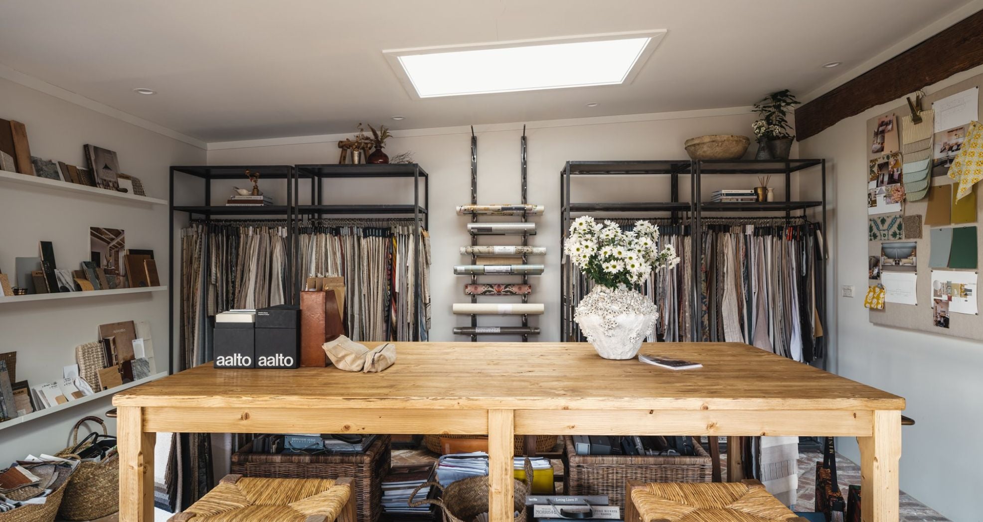 A cozy, well-lit interior design studio with fabric samples hanging on racks and shelves displaying materials. A wooden table with woven chairs is centered, topped with white flowers and design books. Walls show various mood boards and design inspiration.