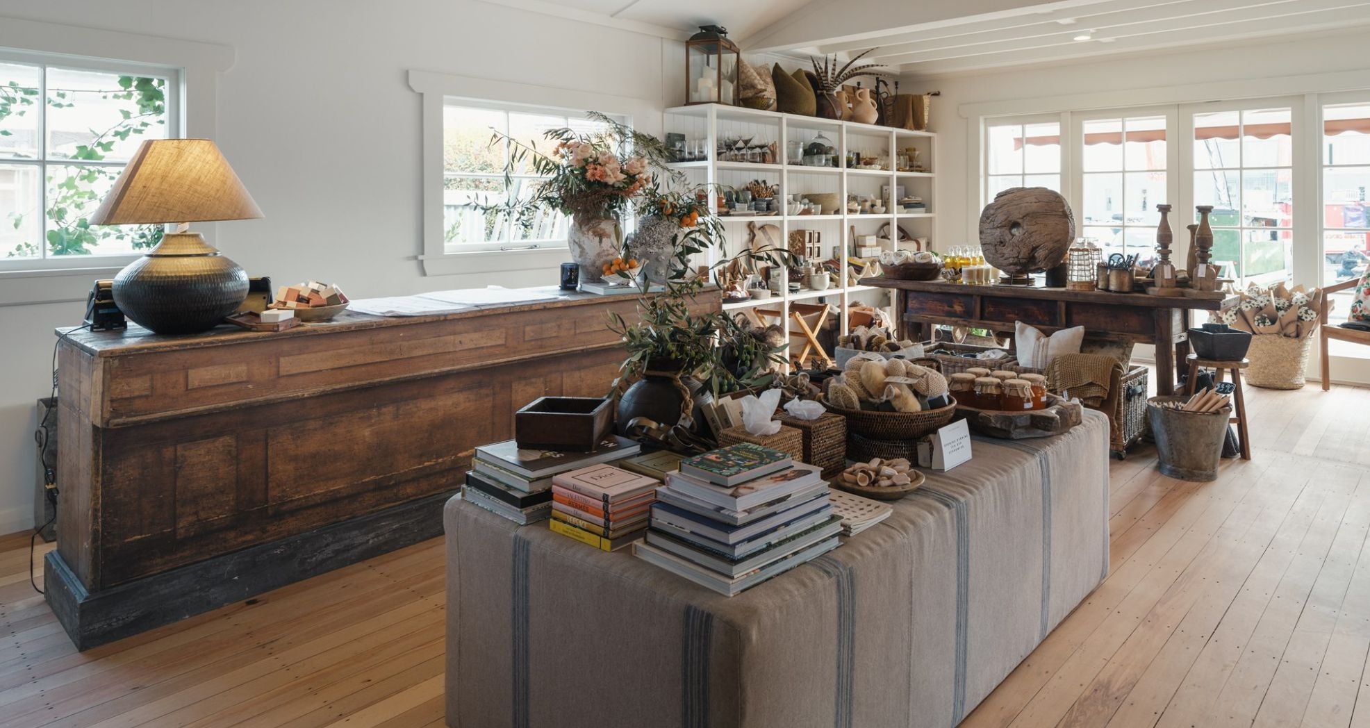 A cozy, rustic store interior with wooden floors and furniture. Displays feature books, candles, and home decor items. Soft lighting and large windows create a warm and inviting atmosphere.