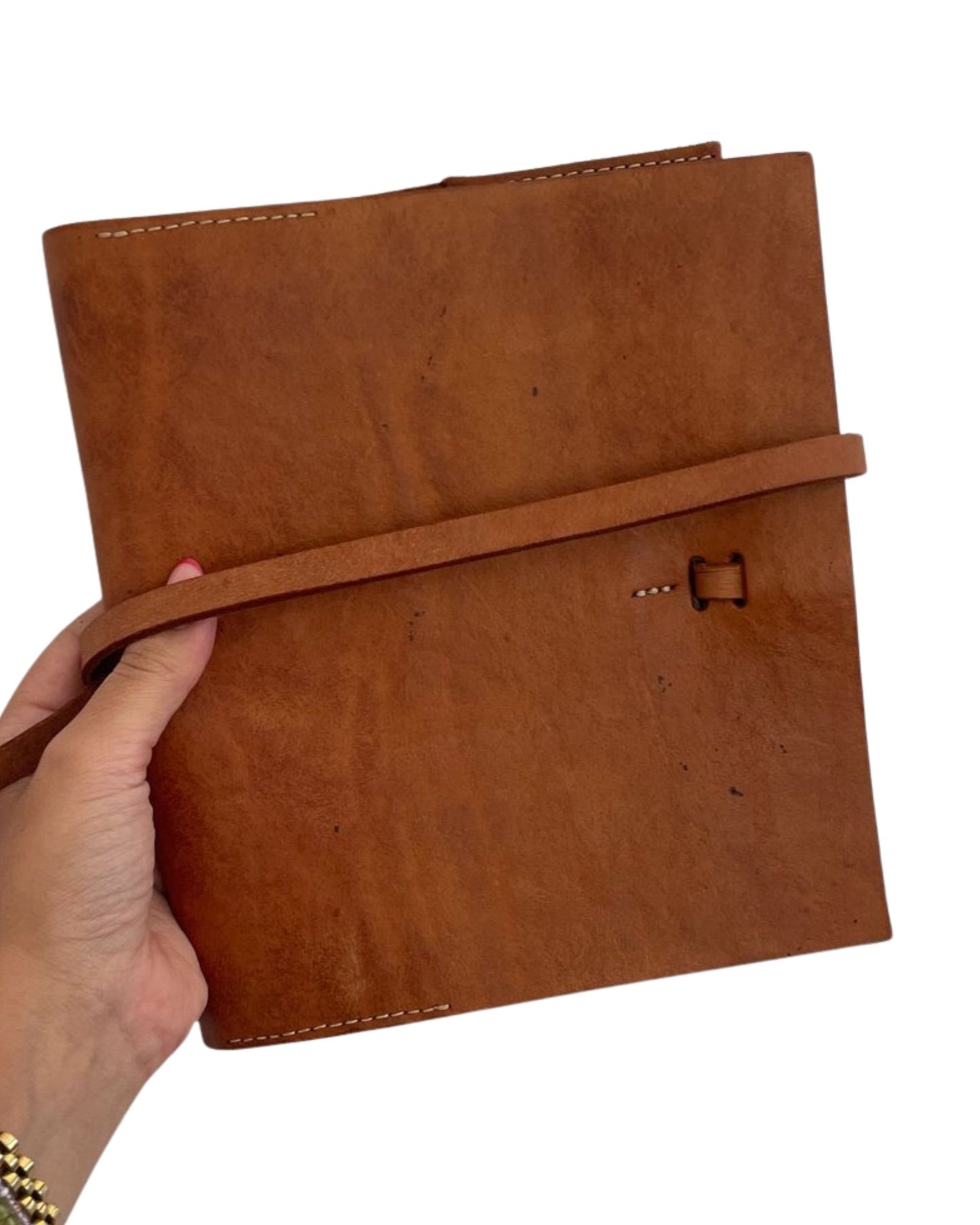 A hand is holding a LEATHER BOOK COVER by The Workroom. It features a brown full-grain leather design with a wrap-around strap, exuding rustic charm. Visible stitching along the edges enhances its timeless appeal.