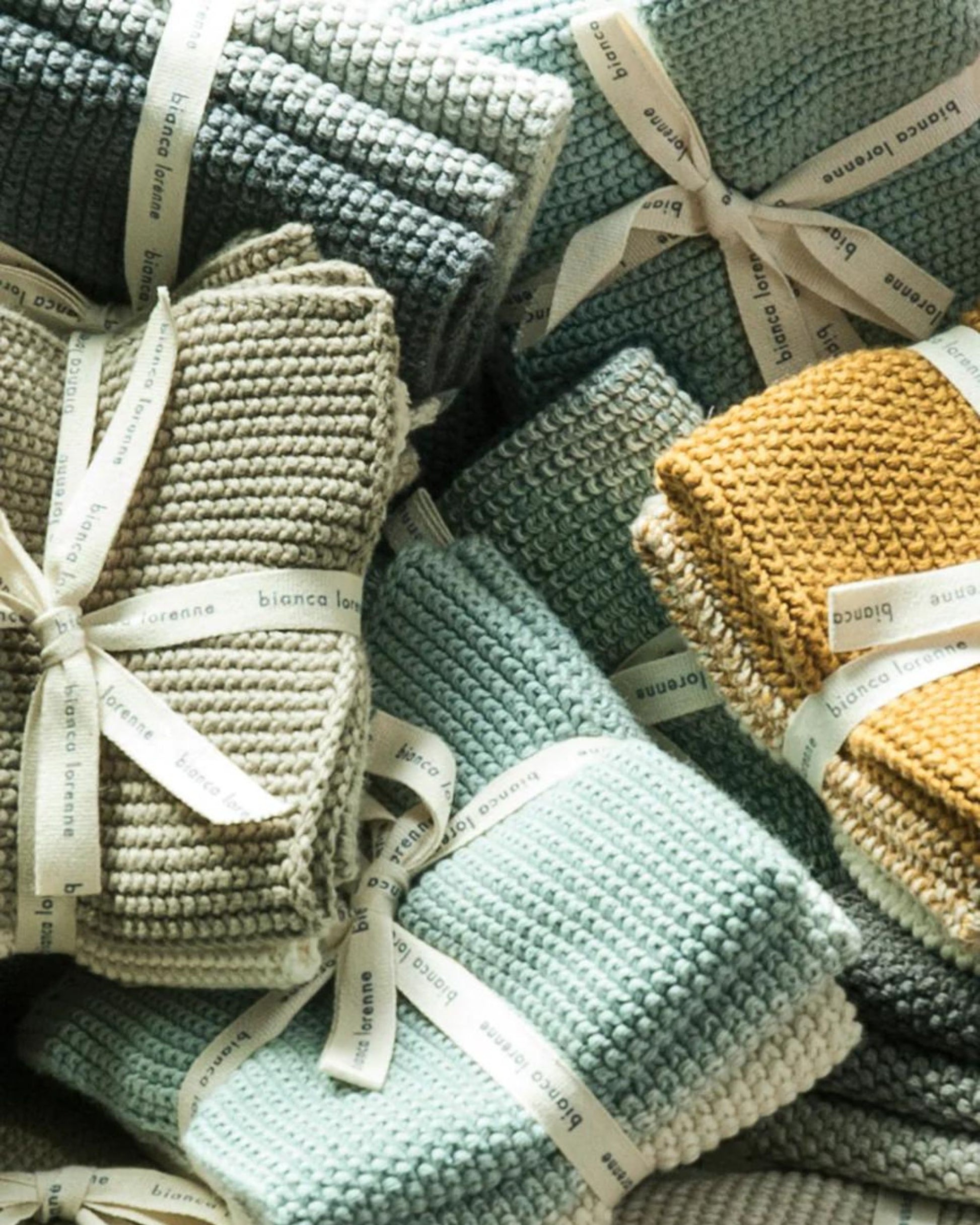 A collection of neatly folded WASH CLOTHS in shades of green, grey, and mustard yellow, each tied with a ribbon labeled Bianca Lorenne. Made from soft organic cotton, their textured surface offers a cozy and artisanal look.