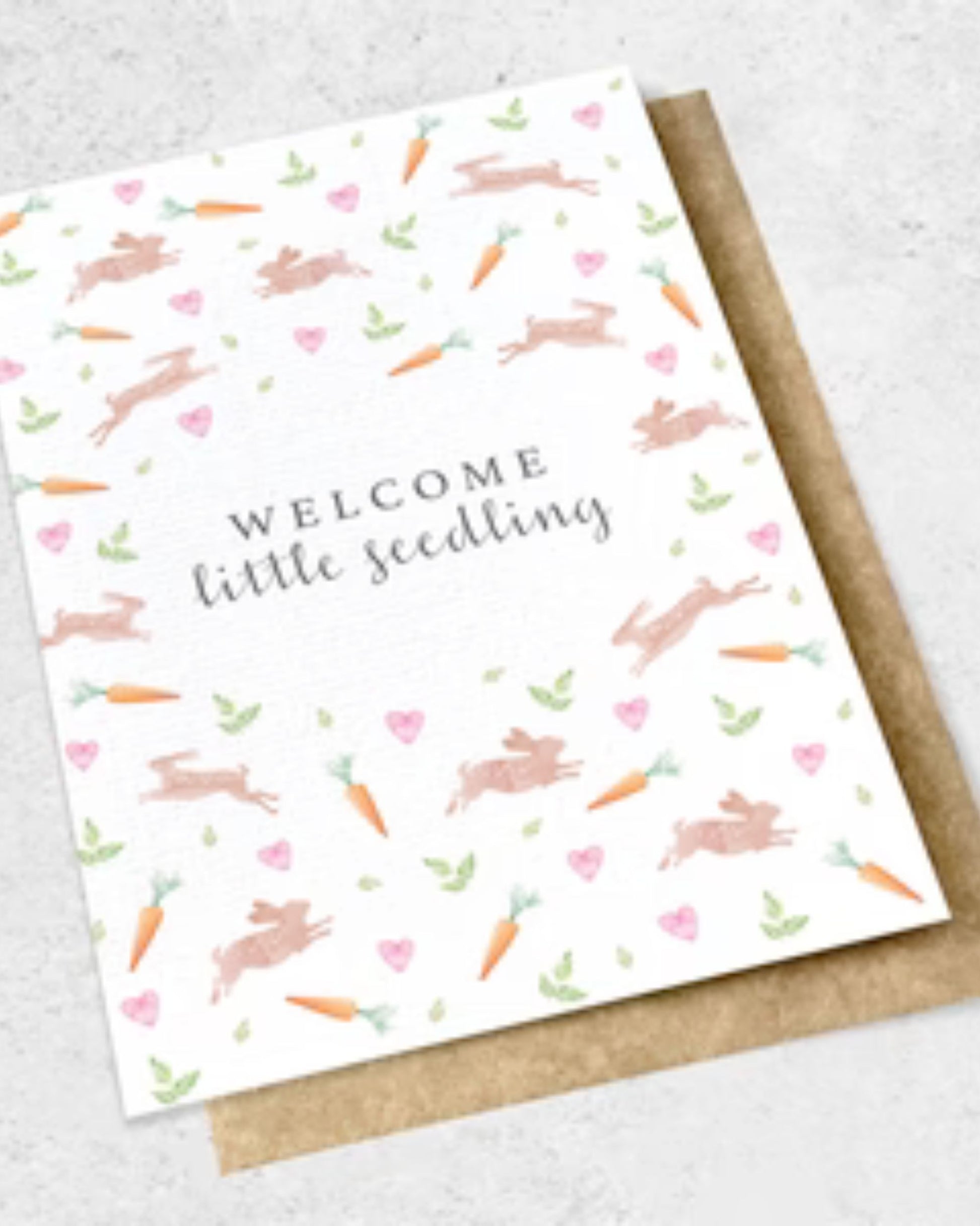 The Ink Bomb WELCOME LITTLE SEEDLING - GREETING CARD features a premium textured design with the phrase "Welcome little seedling" surrounded by charming illustrations of rabbits, carrots, pink hearts, and green leaves. It is set against a kraft envelope on a light gray background.