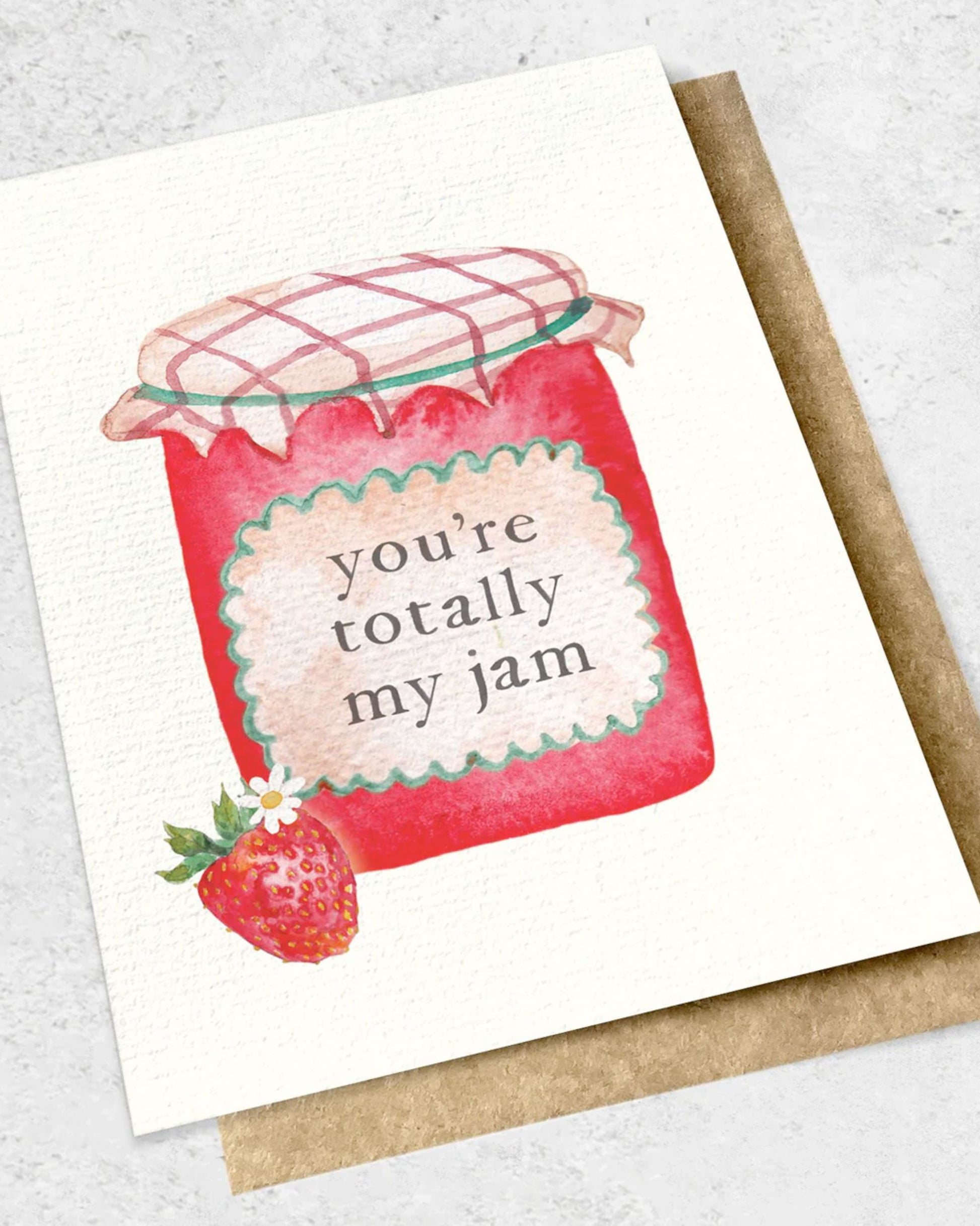 Greeting card titled "YOU'RE TOTALLY MY JAM - GREETING CARD" by Ink Bomb, showcasing a watercolor illustration of a jar with a checkered cloth lid and a label that says "you're totally my jam." A strawberry and small white flower adorn the bottom. It comes with a kraft envelope, all presented on a premium textured surface.