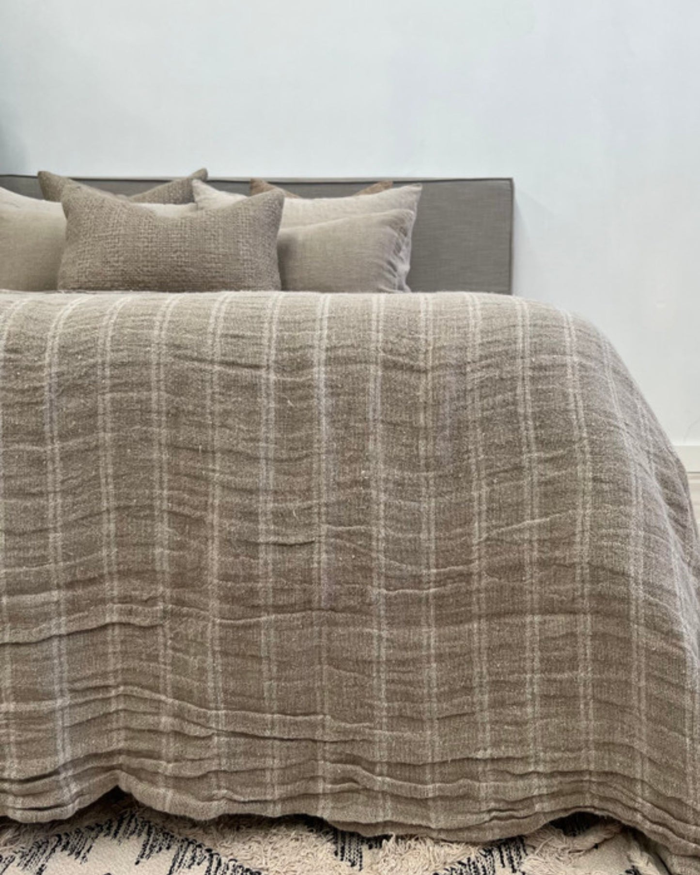 A neatly made bed features the ANGASTON HANDLOOMED LINEN BEDCOVER - WHITE STRIPE by Rustic Linen, adding a handloomed touch with its checkered beige design. Several square throw pillows rest against the gray headboard. The room's white walls and the patterned rug at the bottom complete this serene space.