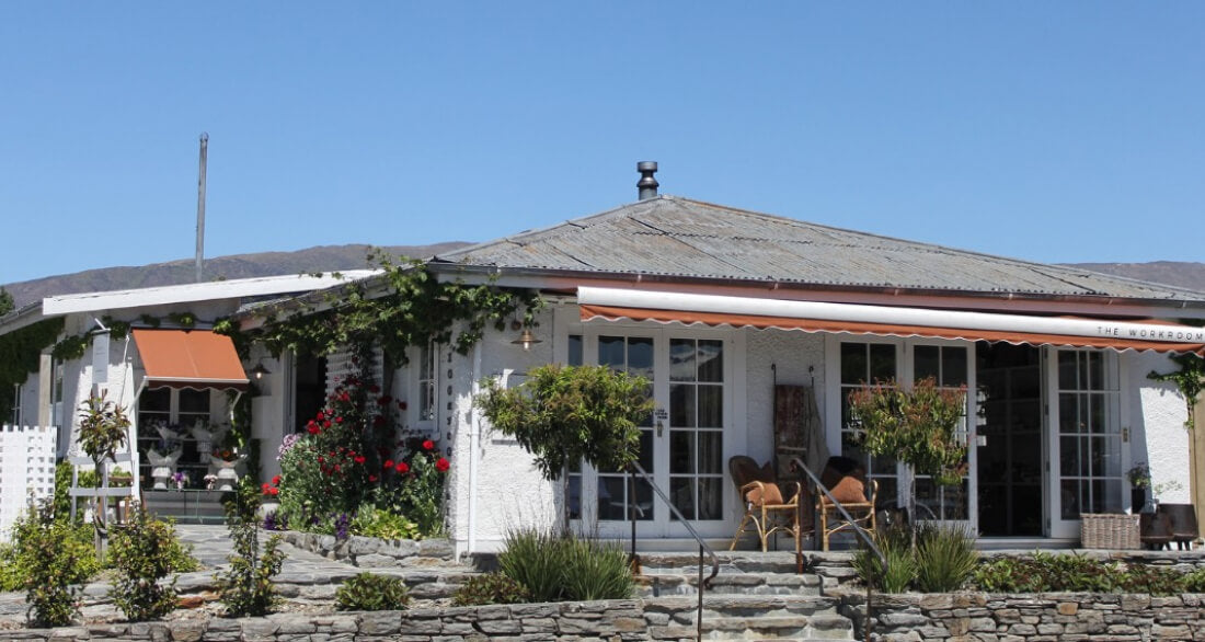 A quaint single-story building with a stone walkway and lush plants. It has a white exterior, wide windows, and a striped awning. Two chairs sit on the porch under the blue sky, creating a welcoming, serene setting.